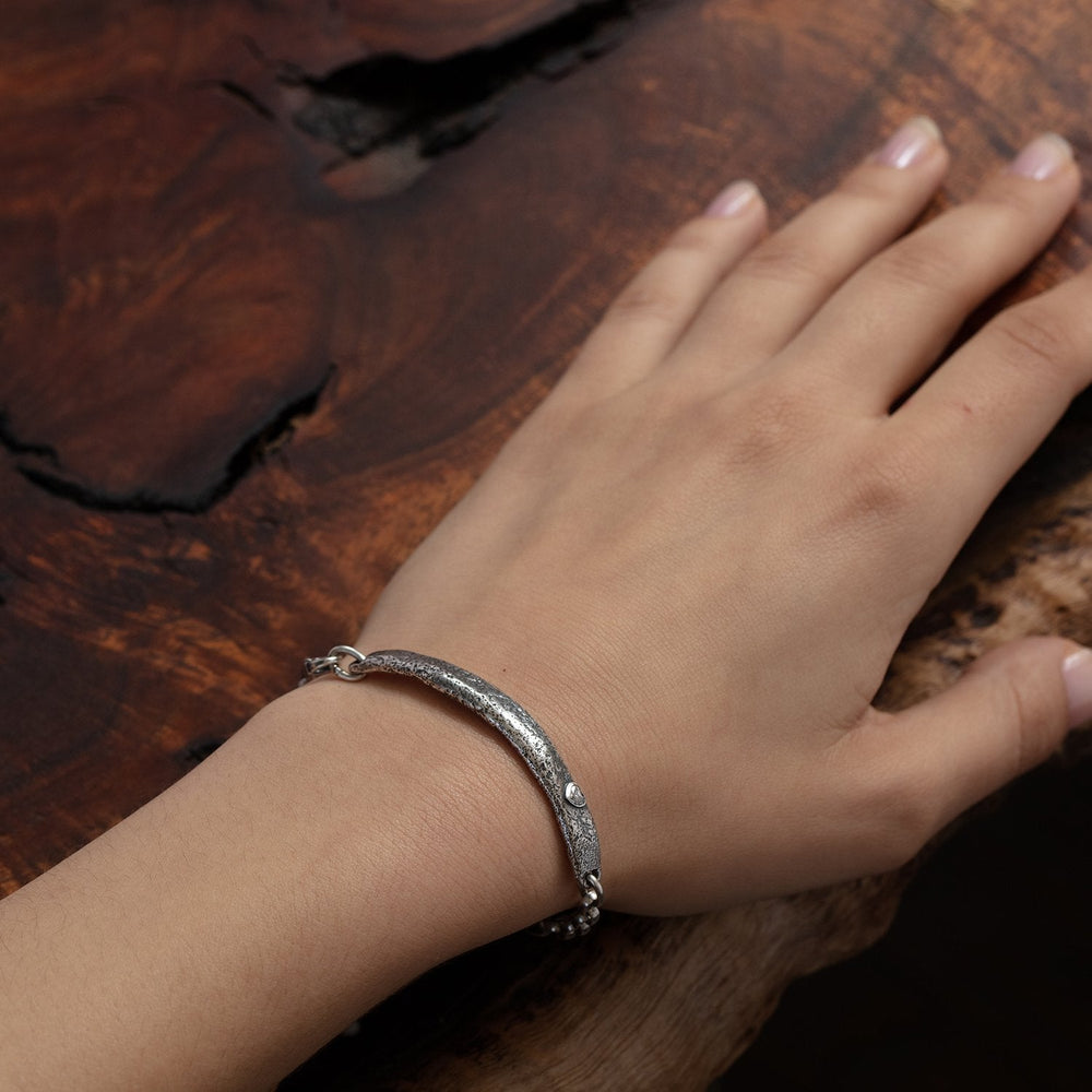 
                      
                        Detail view of model wearing Molten ID Bracelet w/Heart by Betsy Barron on left hand, placed over rough wood table.
                      
                    